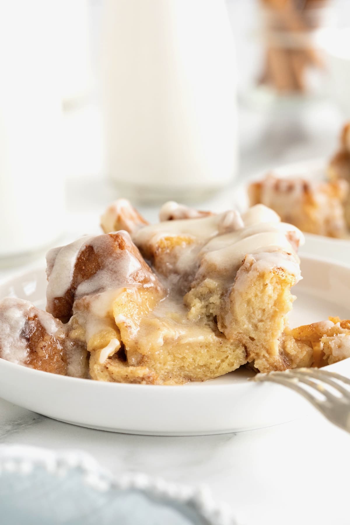 A small white rimmed dessert plate with a serving of bread pudding topped with a white sauce. There is a fork leaning on the plate.