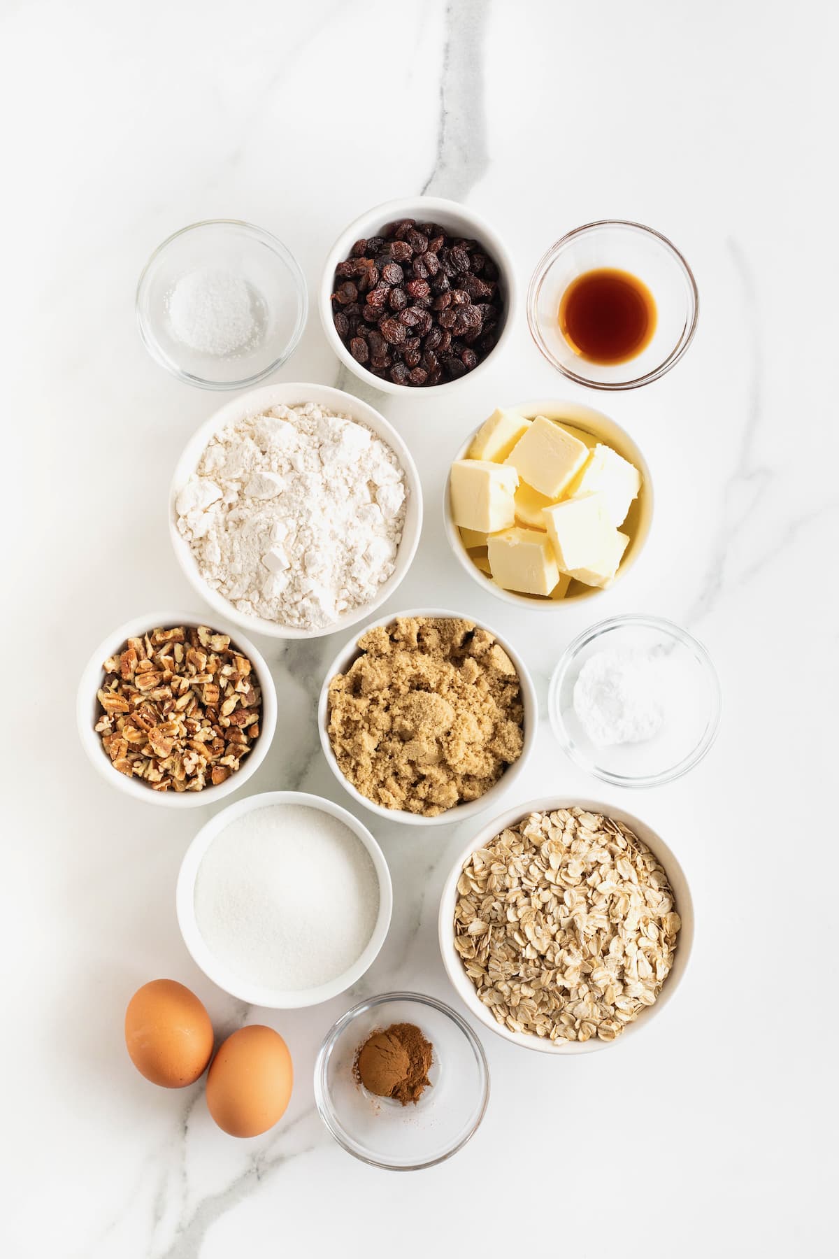 Ingredients to make oatmeal raisin cookies with pecans in small glass dishes on a white marble counter.
