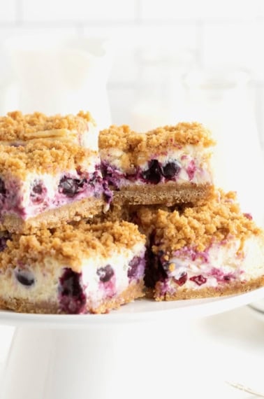 Four lemon blueberry cheesecake bars on a white cake stand.