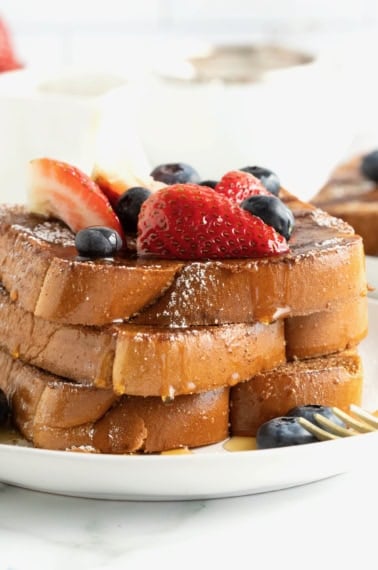 Three slices of baked French toast stacked on a white plate. The French toast is topped with blueberries, strawberry slices and dusted with powdered sugar.