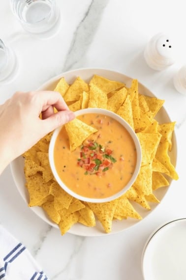 A bowl of queso in the middle of a large white round serving platter filled with corn tortilla chips.