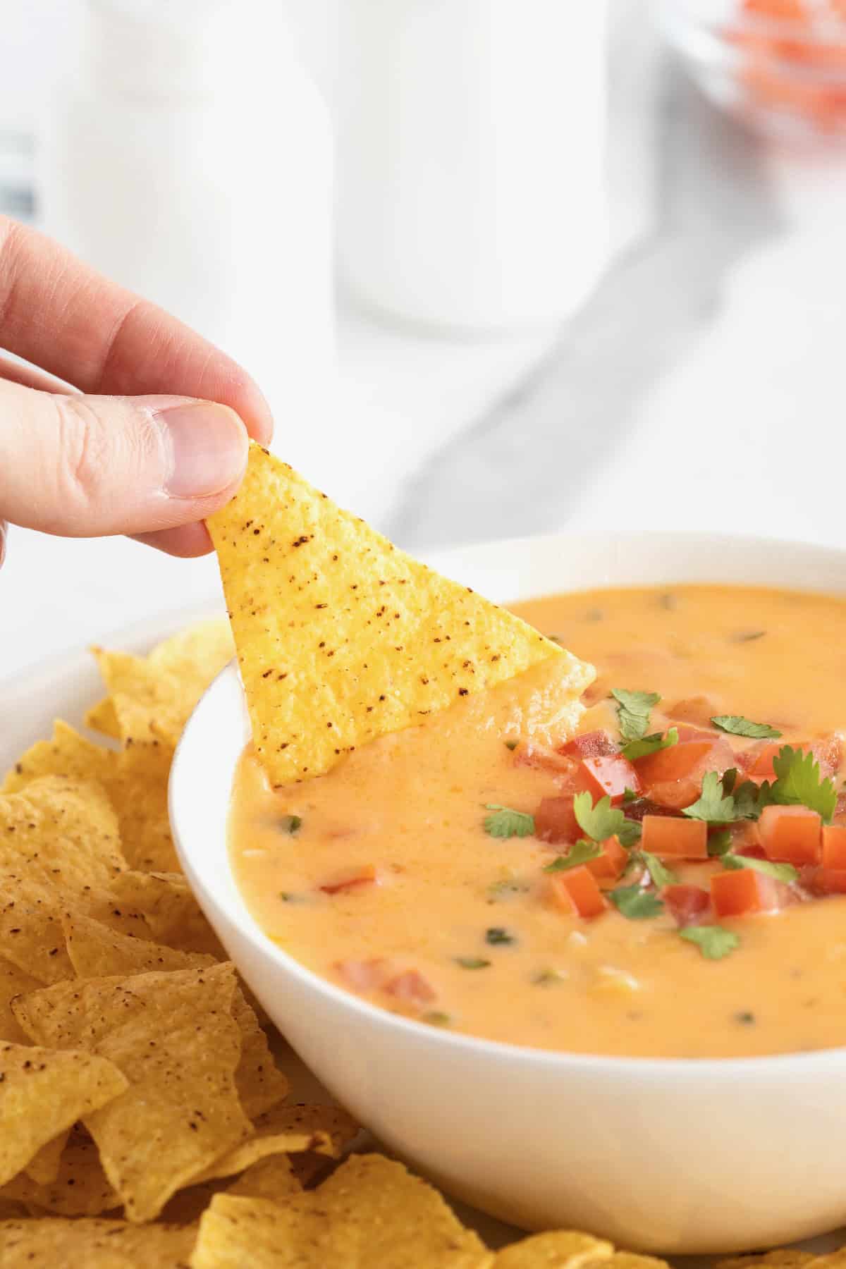 A hand dipping a corn tortilla chip into a white serving bowl of queso.