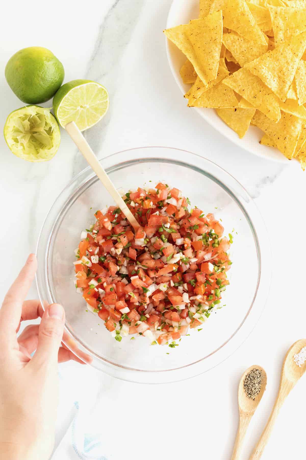 A hand holds the bottom left side of a clear glass mixing bowl of pico de gallo. In the upper right of the photo is a bowl of tortilla chips.  