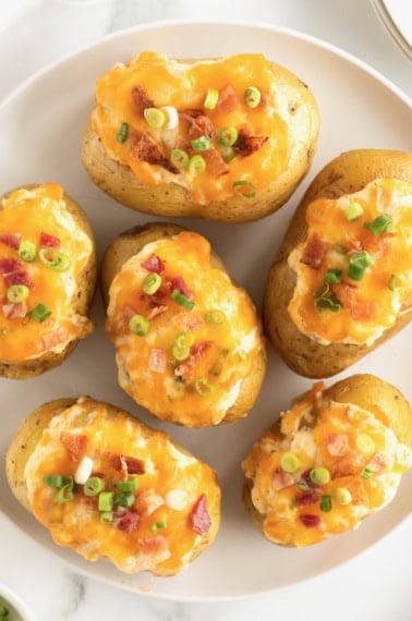 A large white serving plate of twice baked potatoes loaded with bacon pieces, green onion and cheddar cheese.