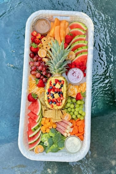 Close up shot of a snack tray floating in a pool. The tray has a halved pineapple in the center and is surrounded by fruits, cheeses and crackers.