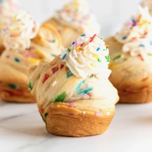 Rainbow sprinkled cruffins topped with simple icing on a white marble counter.