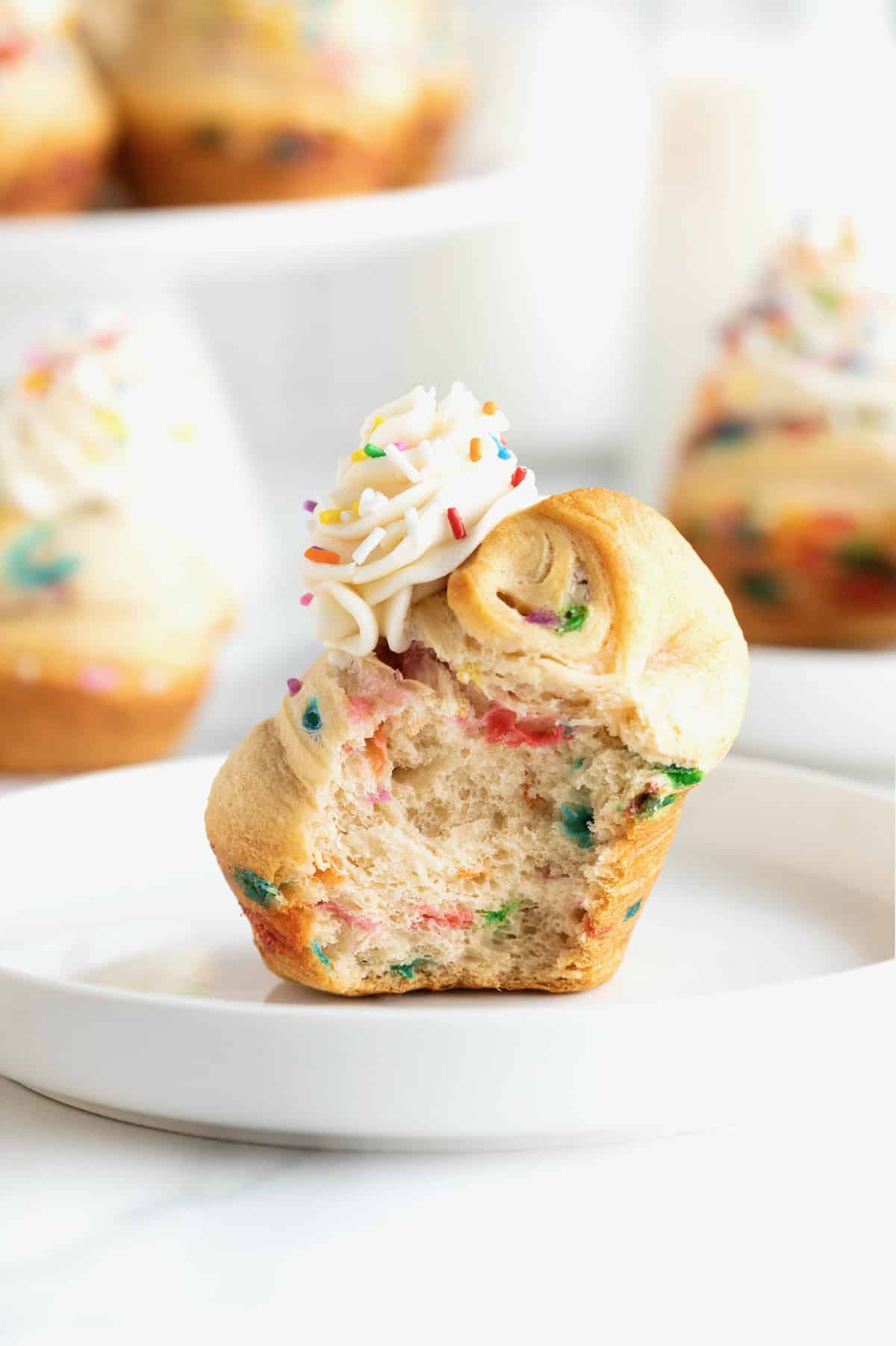 Rainbow sprinkled cruffins topped with simple icing on a white marble counter. There is a bite out of the forward most cruffin.