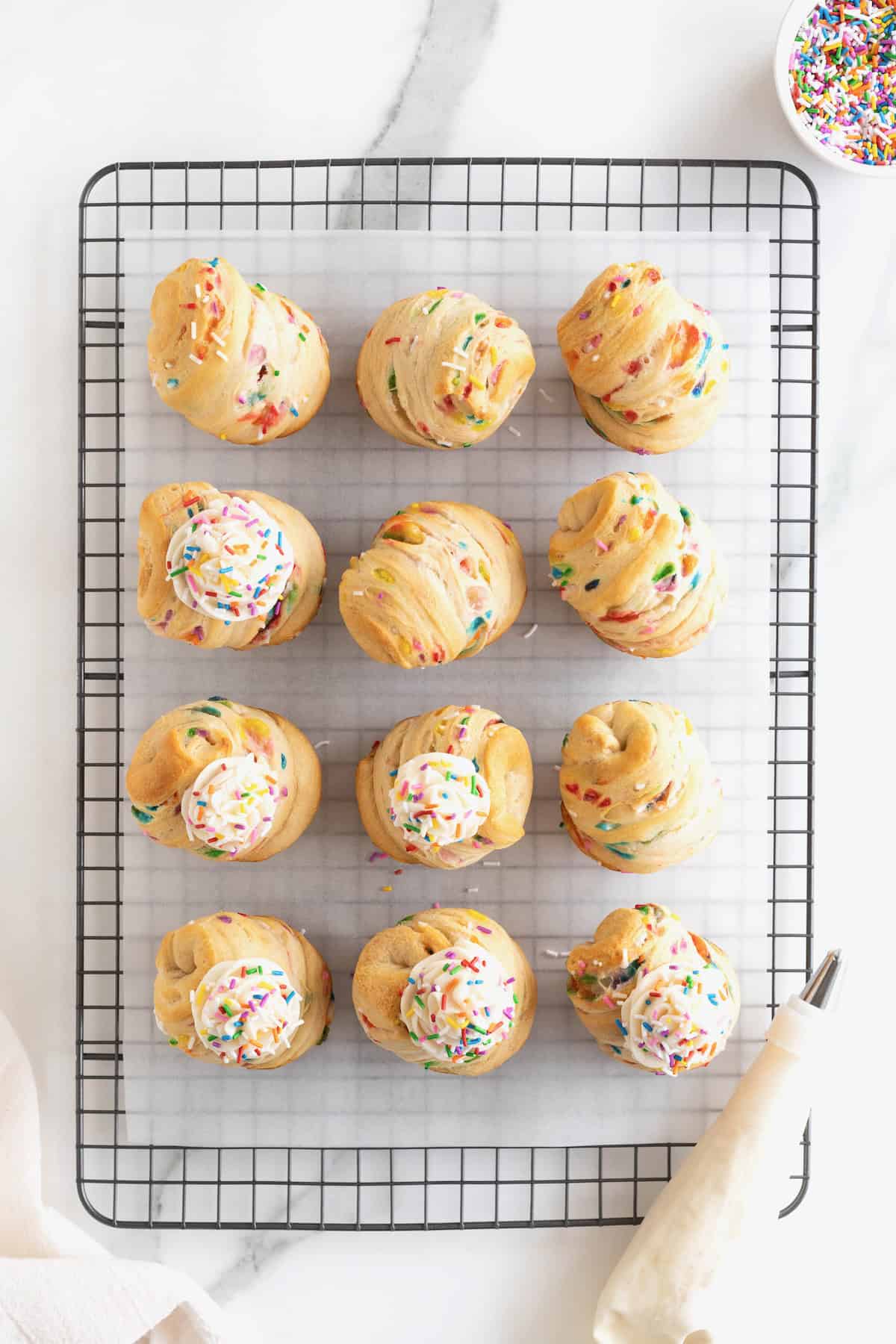 12 cruffins topped with icing and sprinkles on a parchment lined metal cooling rack.