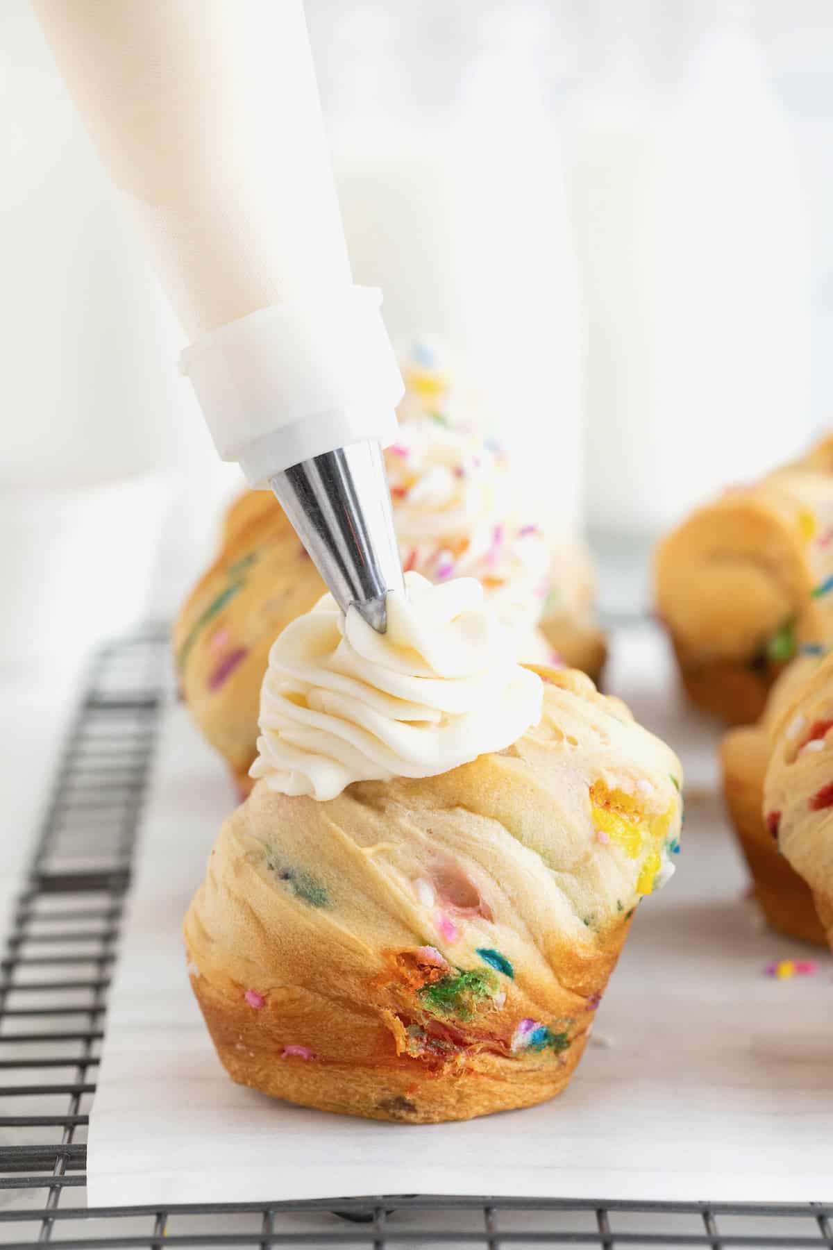 White frosting being piped from a pastry bag onto a confetti cruffin.