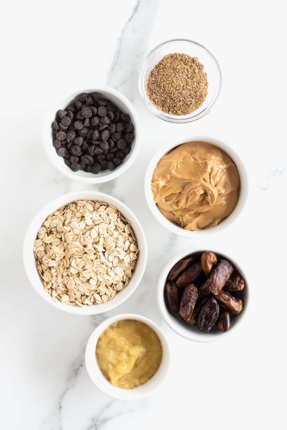 Ingredients to make peanut butter chocolate chip breakfast bars with dates and banana arranged in small white glass dishes on a white marble counter.