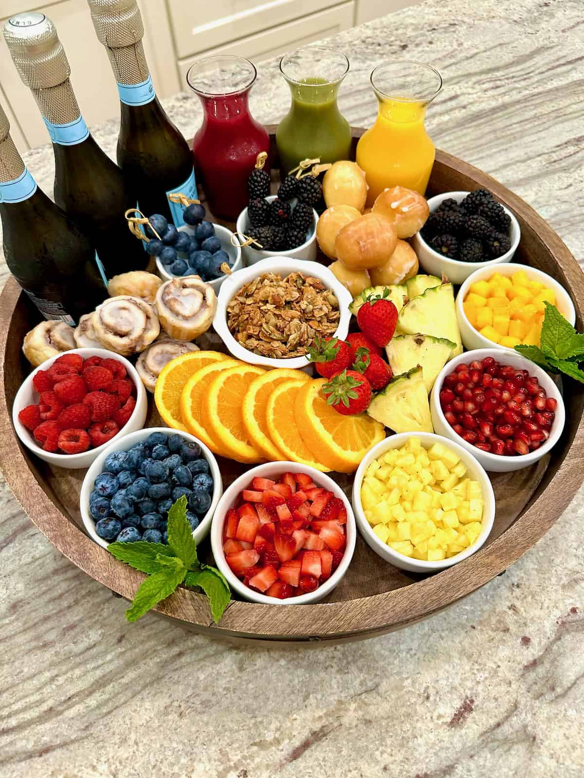A large rimmed wood Lazy Susan filled with colorful fresh fruits in small white dishes, glass carafes of fruit juice and three bottles of Prosecco.