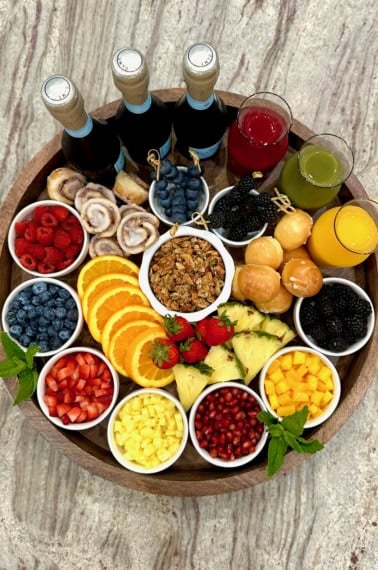 A large round, rimmed wood food board filled with colorful fresh fruits in small white dishes, glass carafes of fruit juice and three bottles of Prosecco.