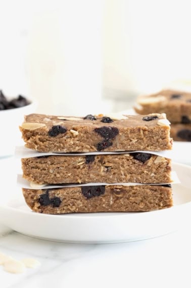 Three blueberry banana almond breakfast bars stacked on a white rimmed plate with pieces of parchment between them.