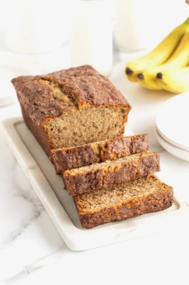 A loaf of banana bread coating in cinnamon sugar on a white stone cutting board on a white marble counter.