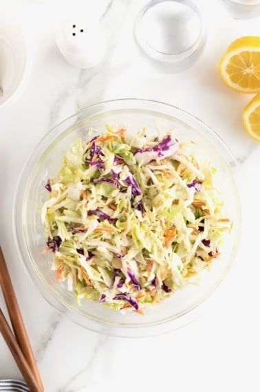 A large glass bowl of coleslaw on a white marble counter. A halved lemon sits to the upper right of the bowl.