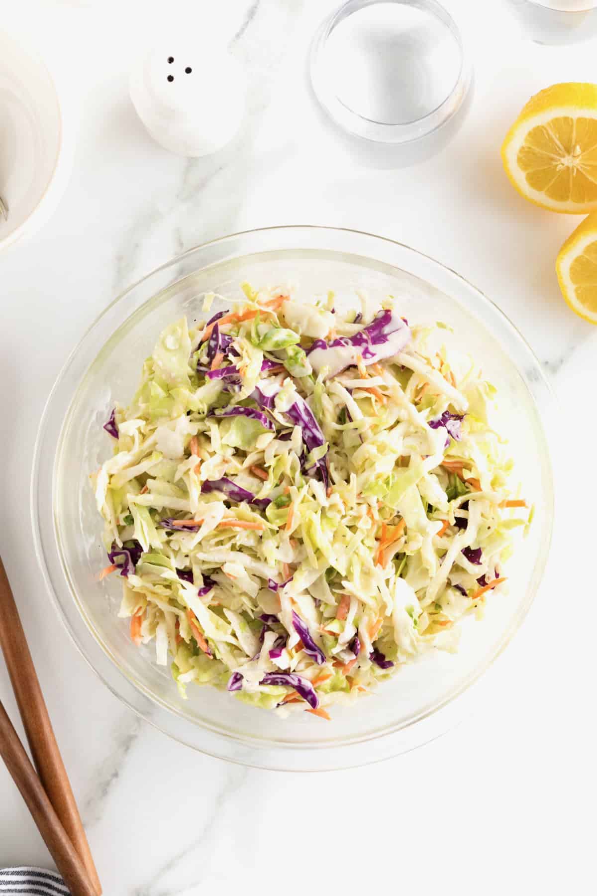 A large glass bowl of coleslaw on a white marble counter. A halved lemon sits to the upper right of the bowl.