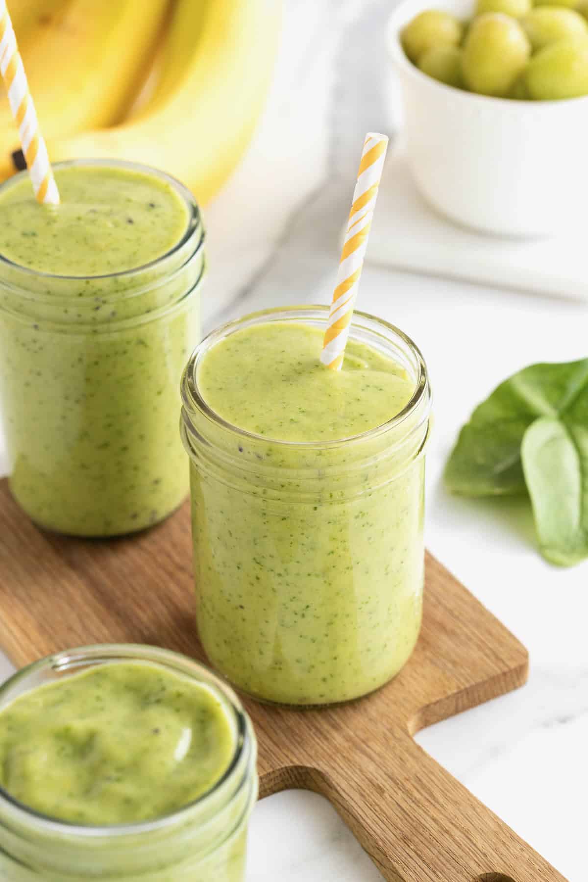 Two mason jars filled with green smoothies sitting on a walnut wood cutting bourdon a white counter. There are yellow and white striped straws in the smoothies.