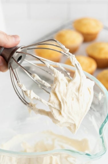 A hand holding the whisk attachment from an electric mixer with white buttercream frosting on it.