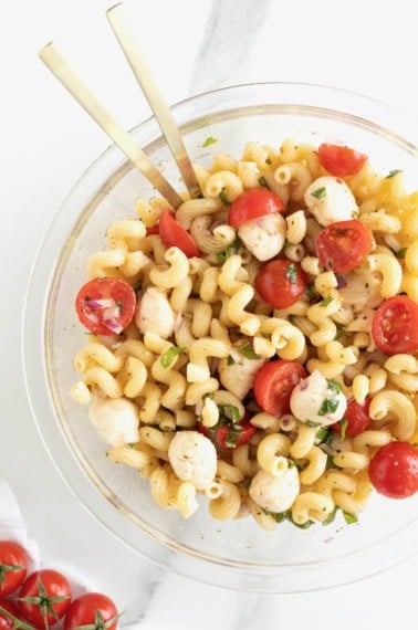 A large glass serving bowl of capers pasta salad with halved cheery tomatoes, bite sized mozzarella balls and corkscrew pasta.