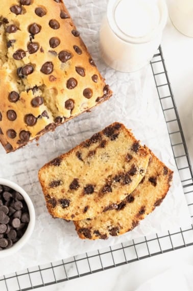 A slice of chocolate chip pound cake in front of a loaf of pound cake on a parchment lined cooling rack. A small white dish of chocolate chips sits to the bottom left of the photo.