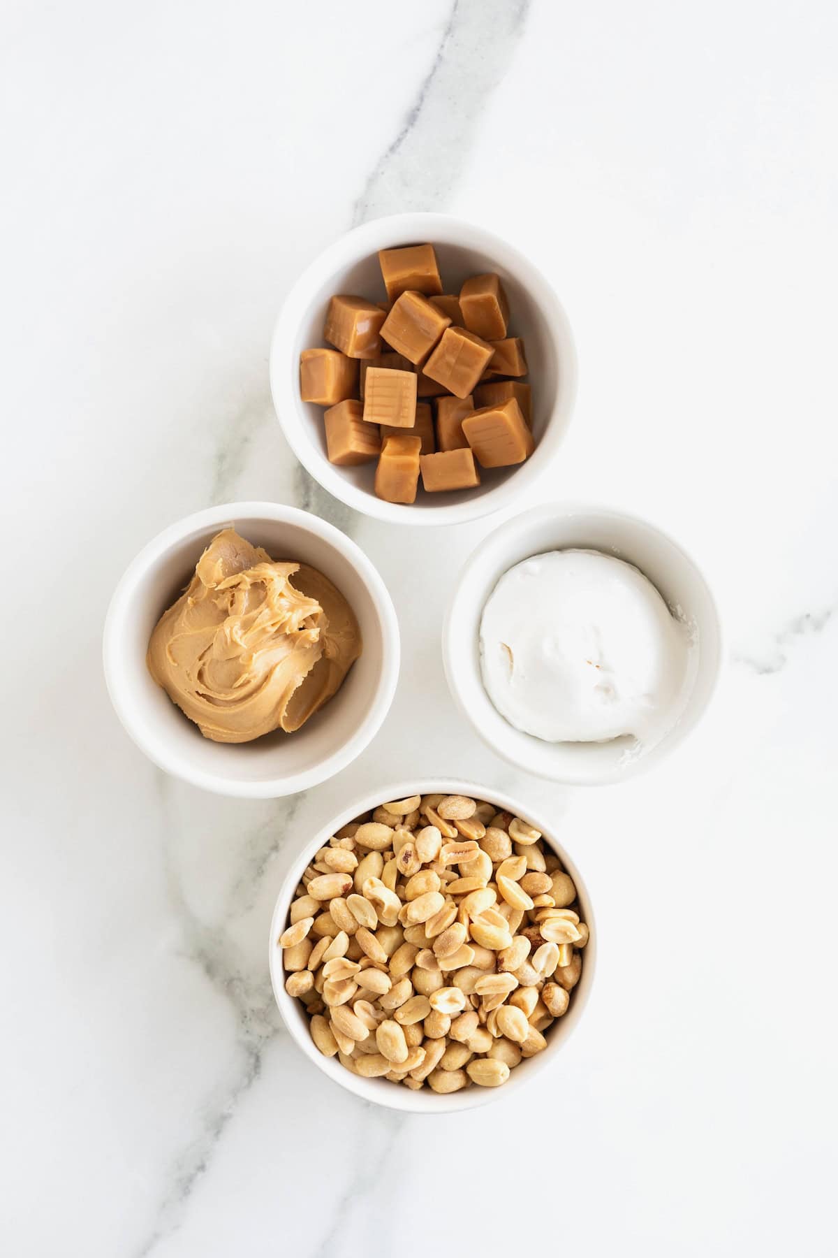 Caramel squares, peanut butter, marshmallow fluff, and peanuts in small white dishes on a white marble counter.