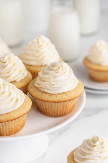 Four vanilla cupcakes with white icing on a white cake stand.