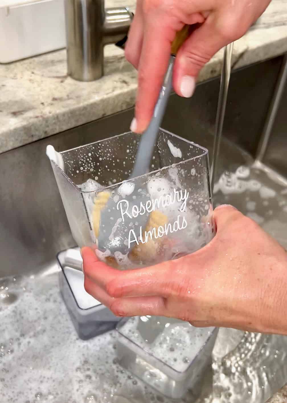 A hand holding a scrub brush, cleaning a clear container in a sink.