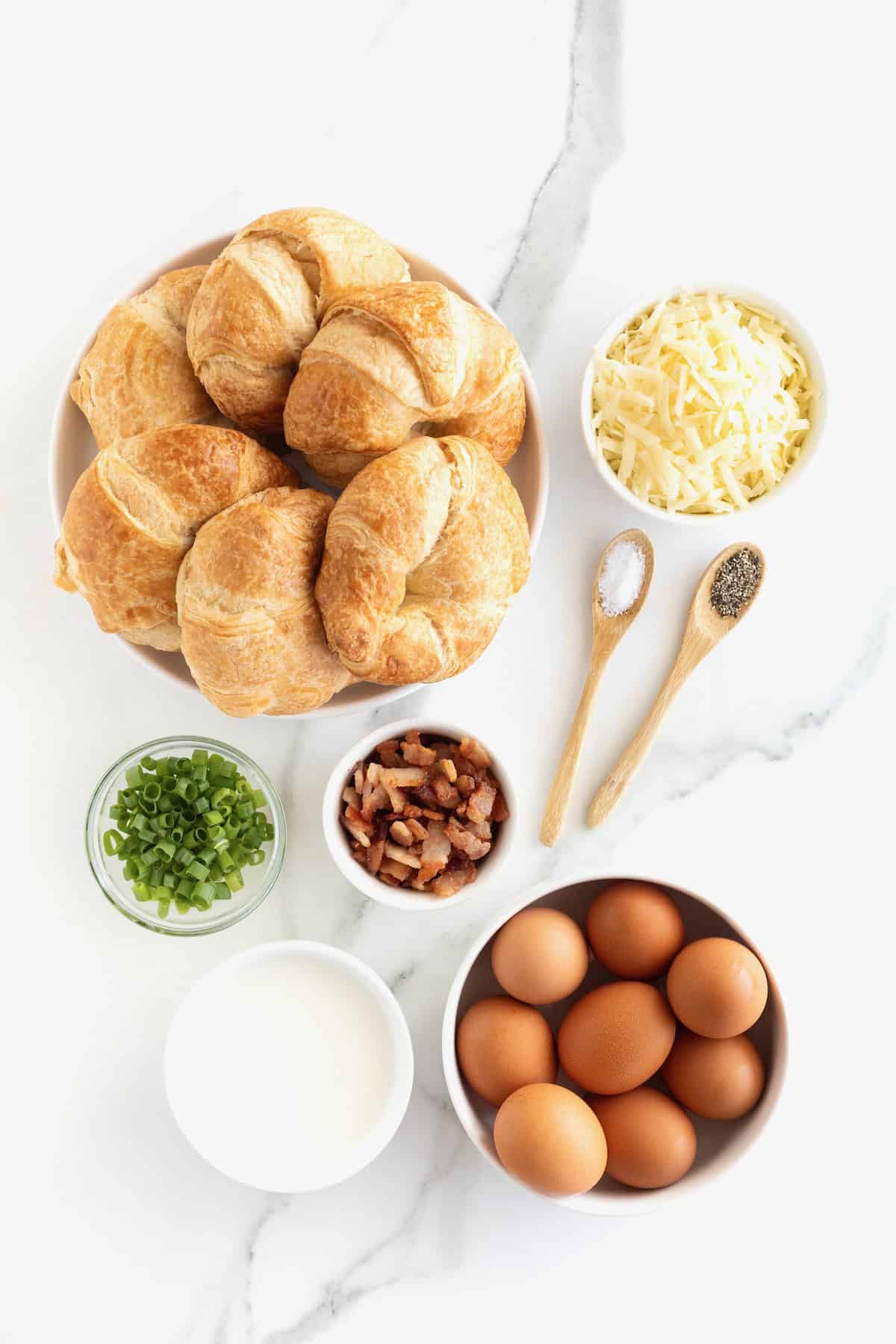 Ingredients to make croissant breakfast casserole in glass dishes on a white marble counter.