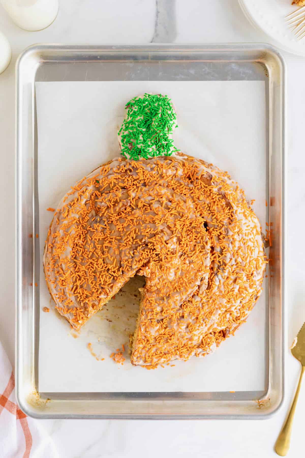 A giant pumpkin-shaped cinnamon roll on a parchment lined aluminum baking sheet. One slice is missing from the cinnamon roll.