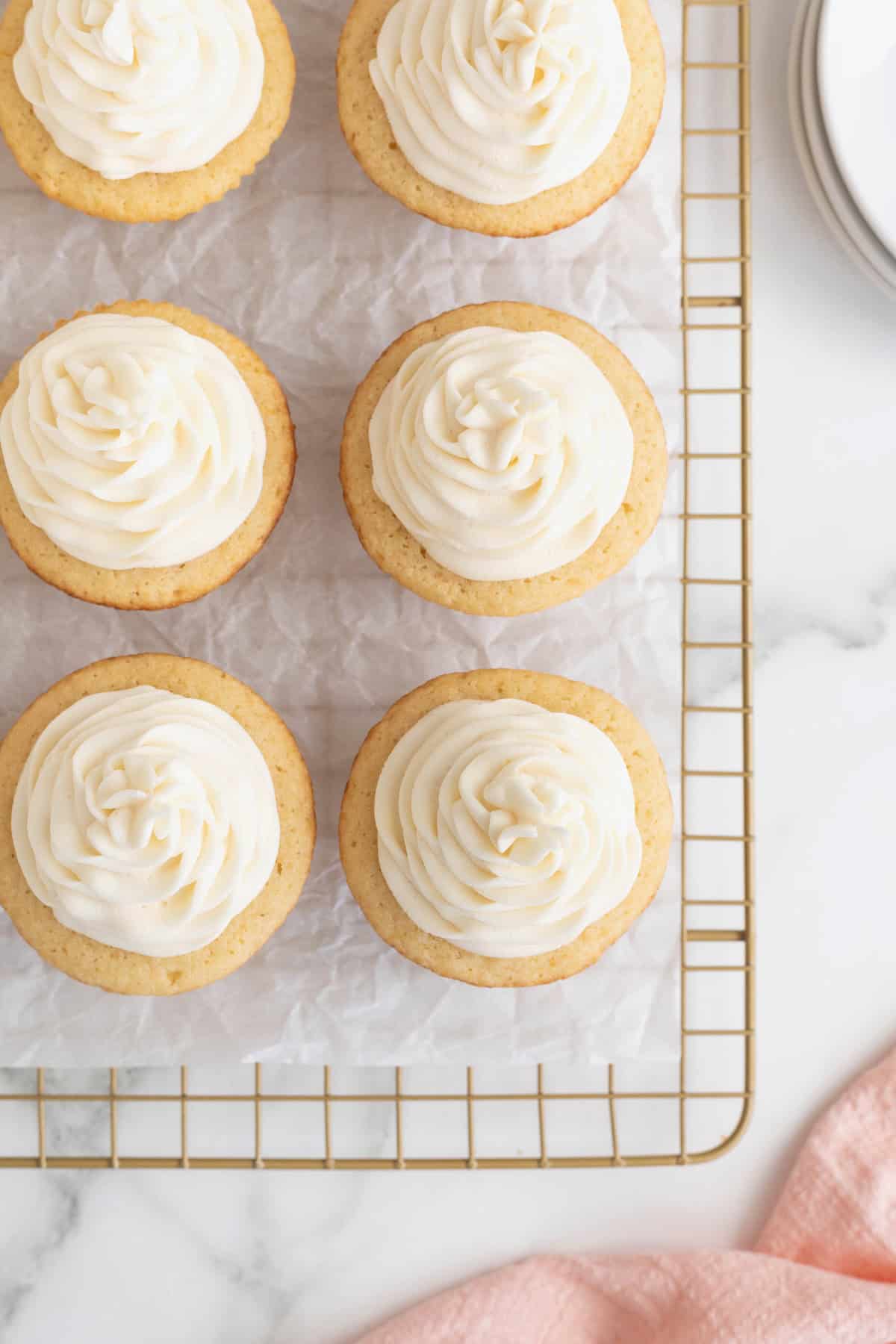 Six vanilla frosted cupcakes on a parchment lined cooling rack. 