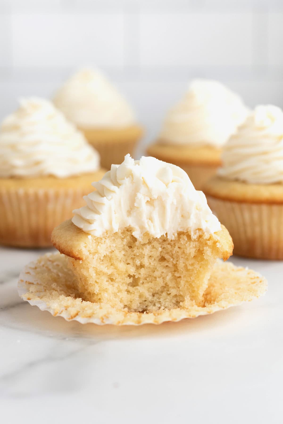 A frosted vanilla cupcake with the paper peeled back and a bite taken out o fit. 