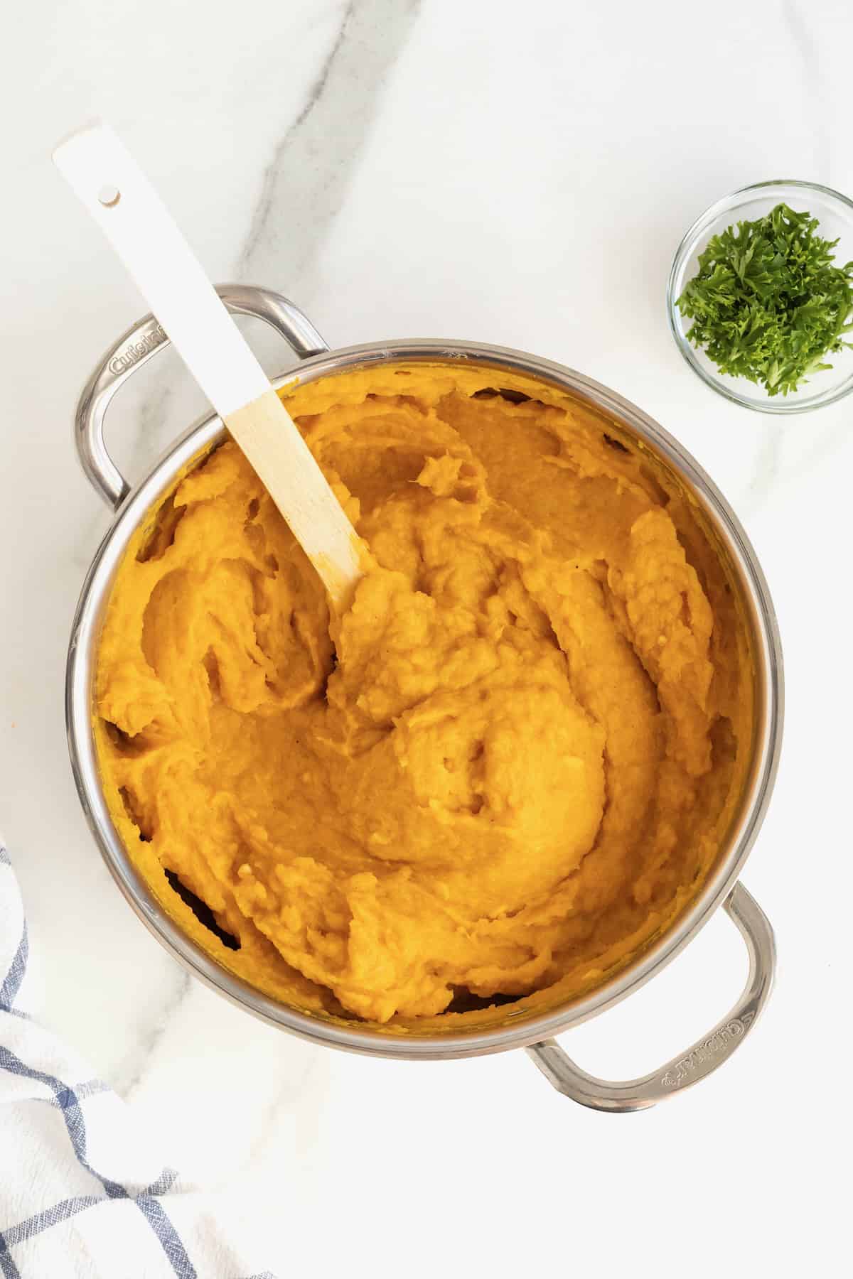 A stainless steel sauce pan of mashed butternut squash on a white marble counter. In the pot is a wooden spoon.