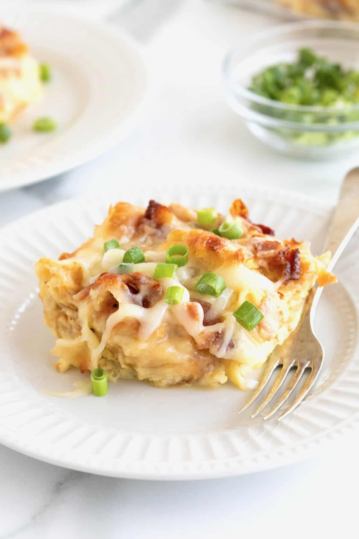 A slice of croissant breakfast casserole on a white fluted plate with a metal fork.