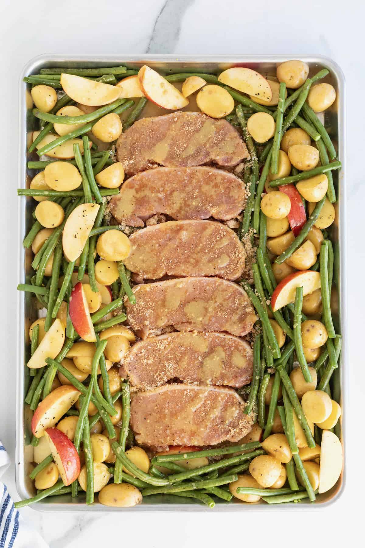 Six pork chops on a rimmed baking sheet surrounded by green beans, apples, and baby potatoes.