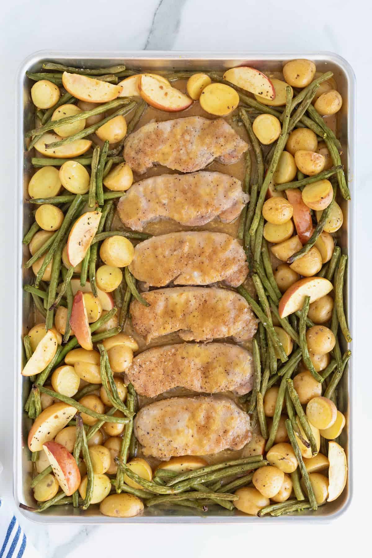 Six pork chops on a rimmed baking sheet surrounded by green beans, apples, and baby potatoes.