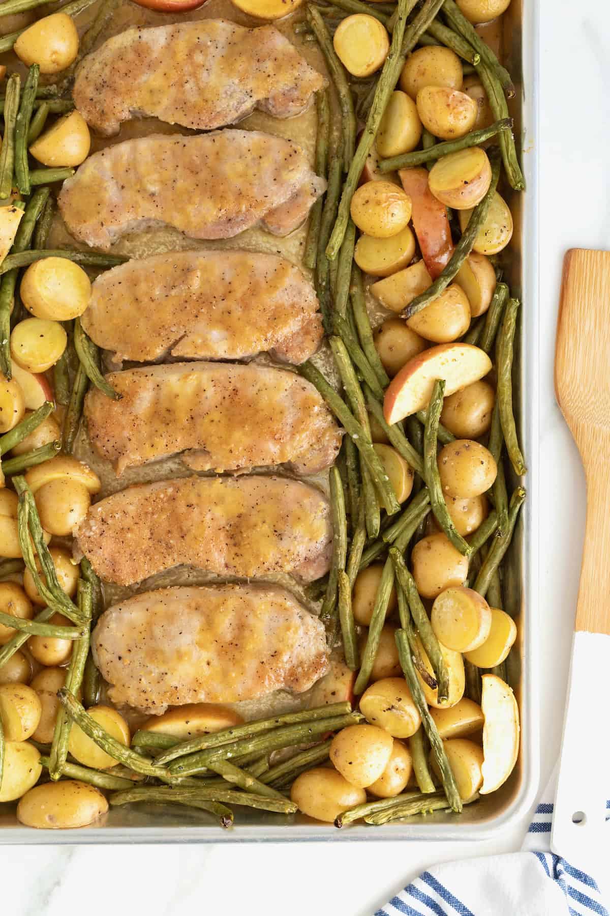 Six pork chops on a rimmed baking sheet surrounded by green beans, apples, and baby potatoes.