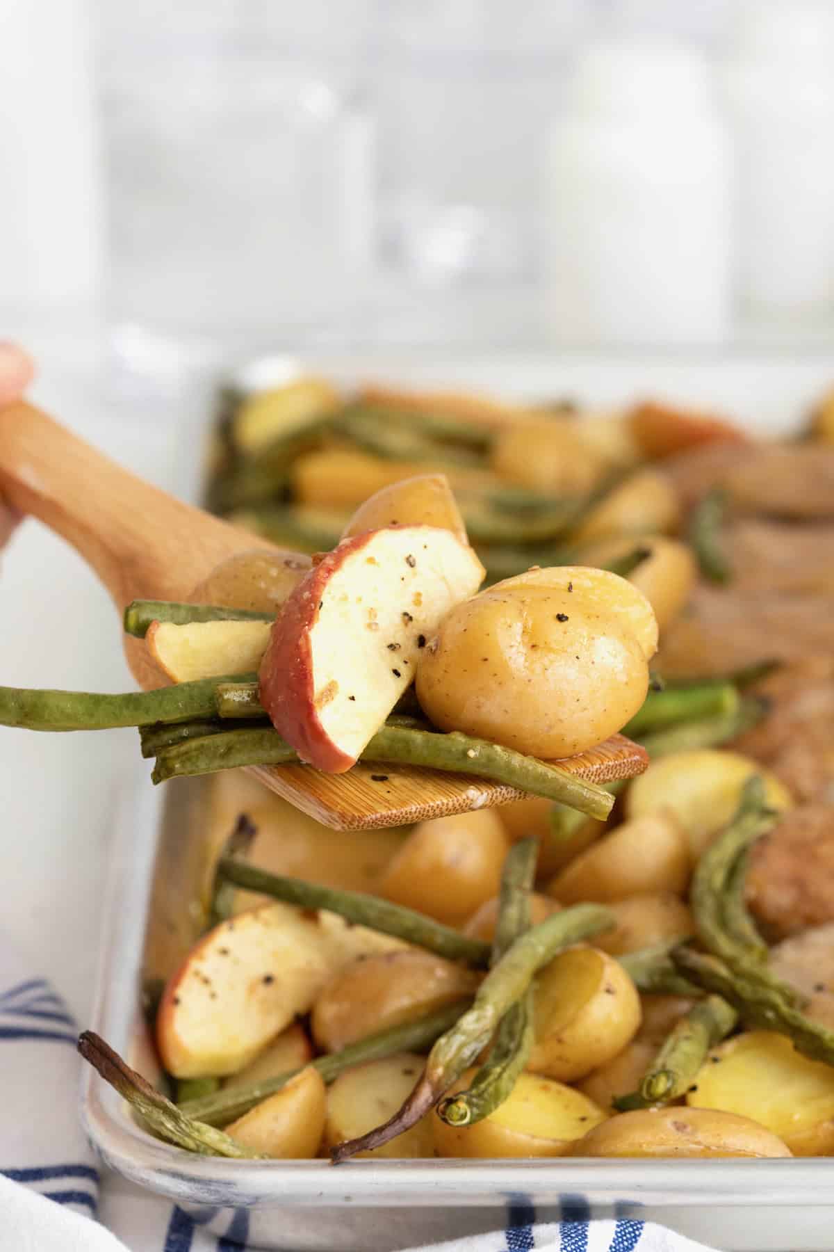 A wooden spatula serving up sheet pan pork chops with green beans, apples and potatoes.