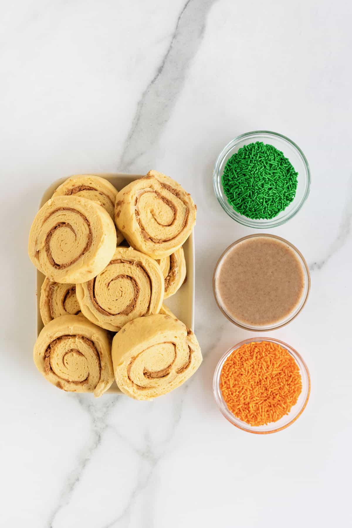 Ingredients to make a giant cinnamon roll pumpkin in small glass dishes on a white marble counter.