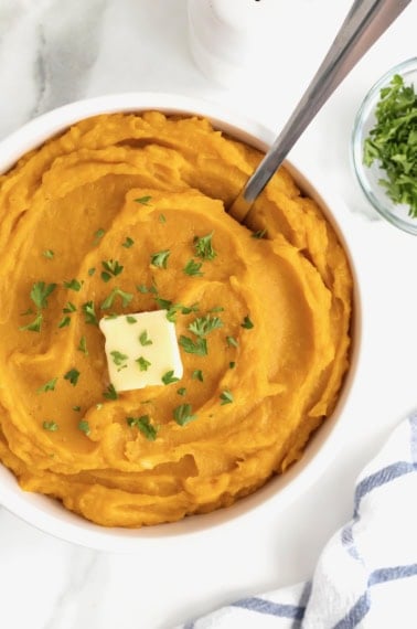 A large white serving bowl of mashed butternut squash topped with a square of butter and fresh parsley.