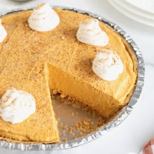 A pumpkin cream pie in a disposable pie plate topped with dollops of whipped cream. There is one piece missing from the pie.