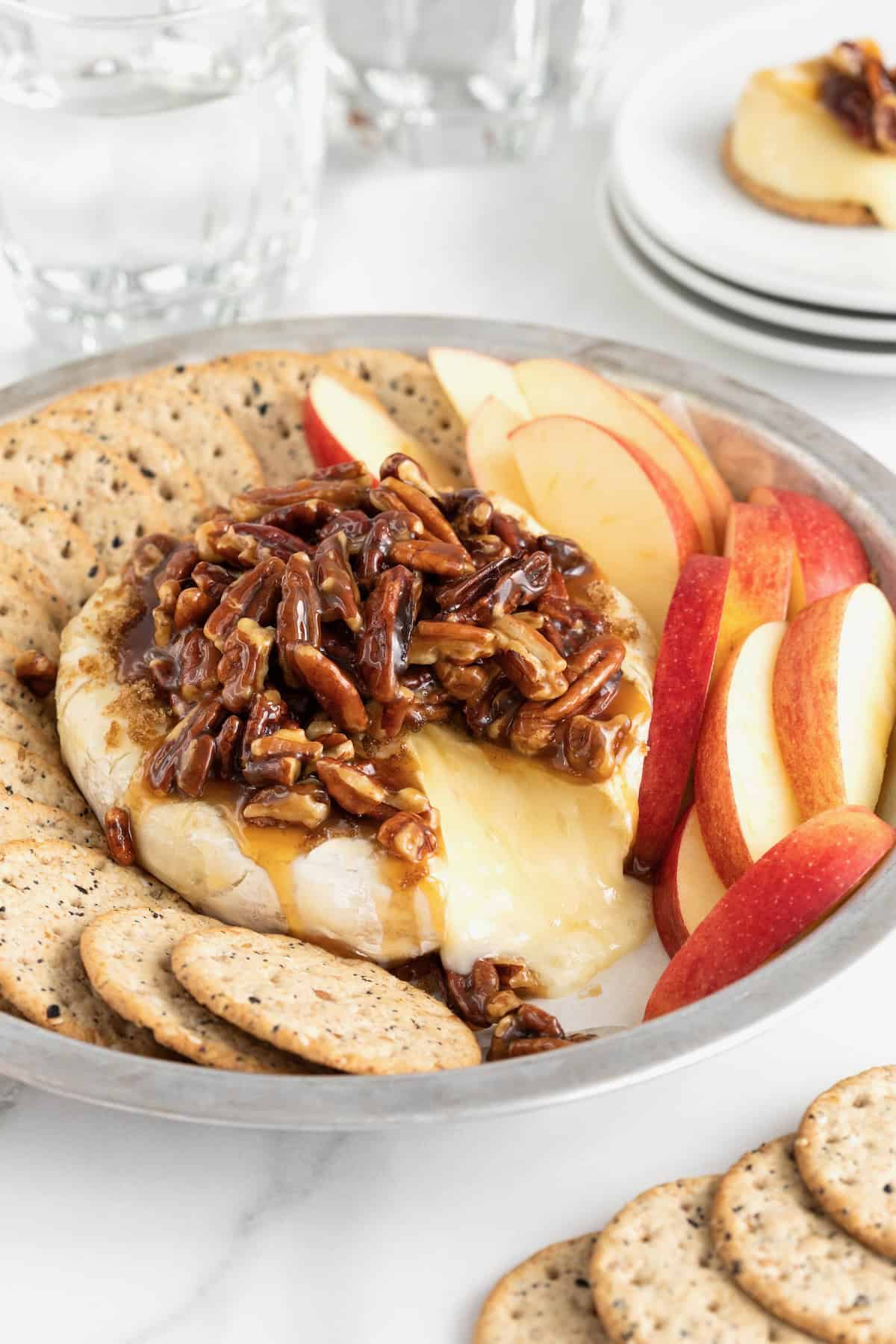 A round aluminum baking dish with a wheel of brie in the middle, and apple slices and water crackers around the outside. The brie is topped with a pecan sauce.