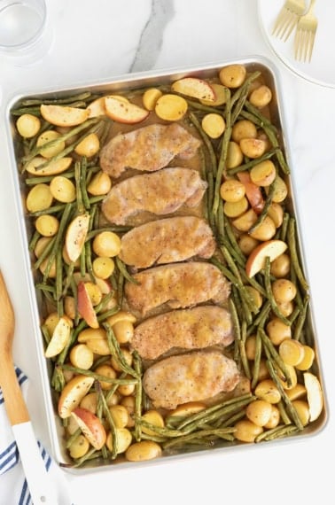 Six pork chops on a rimmed baking sheet surrounded by green beans, apples, and baby potatoes.