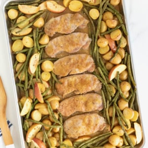 Six pork chops on a rimmed baking sheet surrounded by green beans, apples, and baby potatoes.