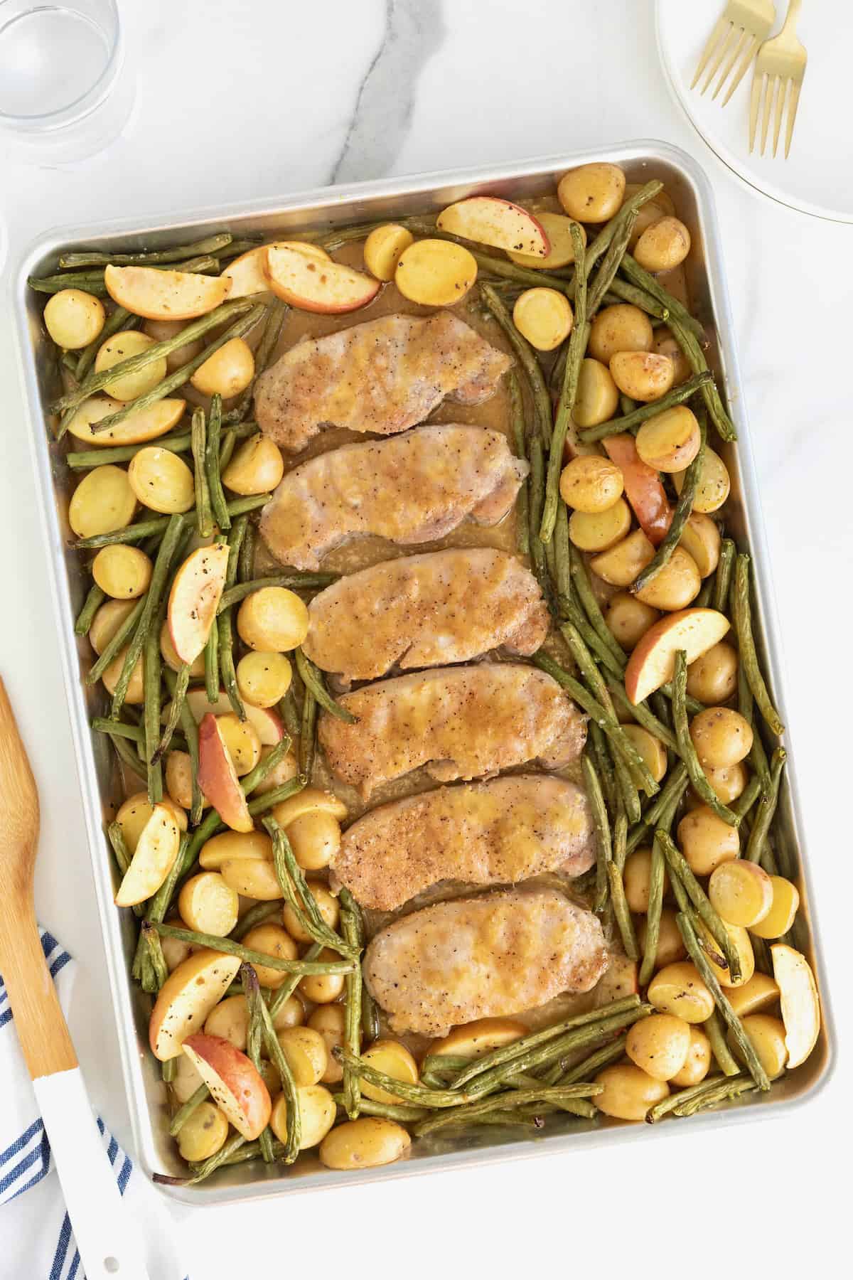 Six pork chops on a rimmed baking sheet surrounded by green beans, apples, and baby potatoes.