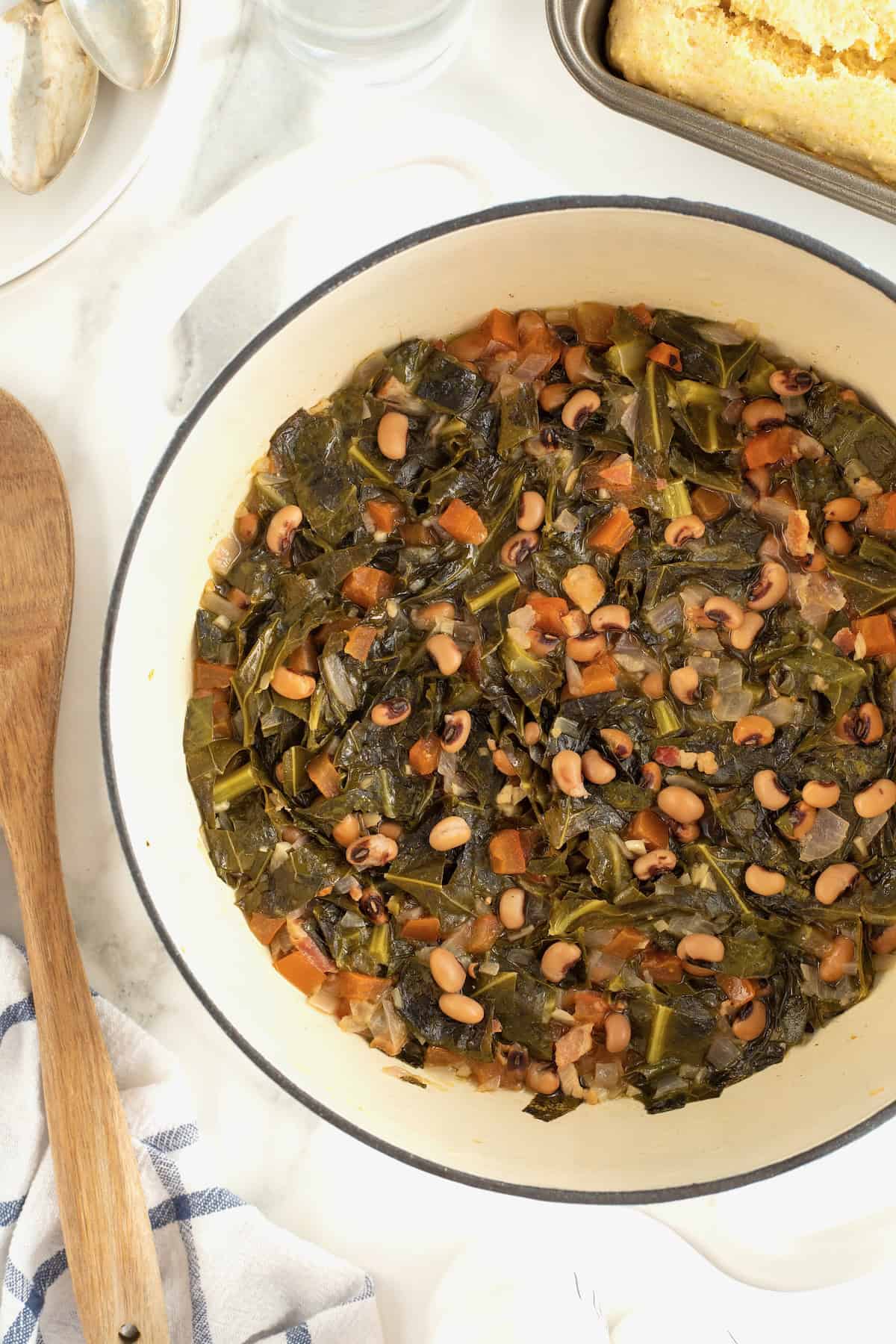 A cream colored dutch oven filled with black eyed peas and collard greens on a white marble counter. Next to the Dutch oven on the left hand side is a wooden spoon.