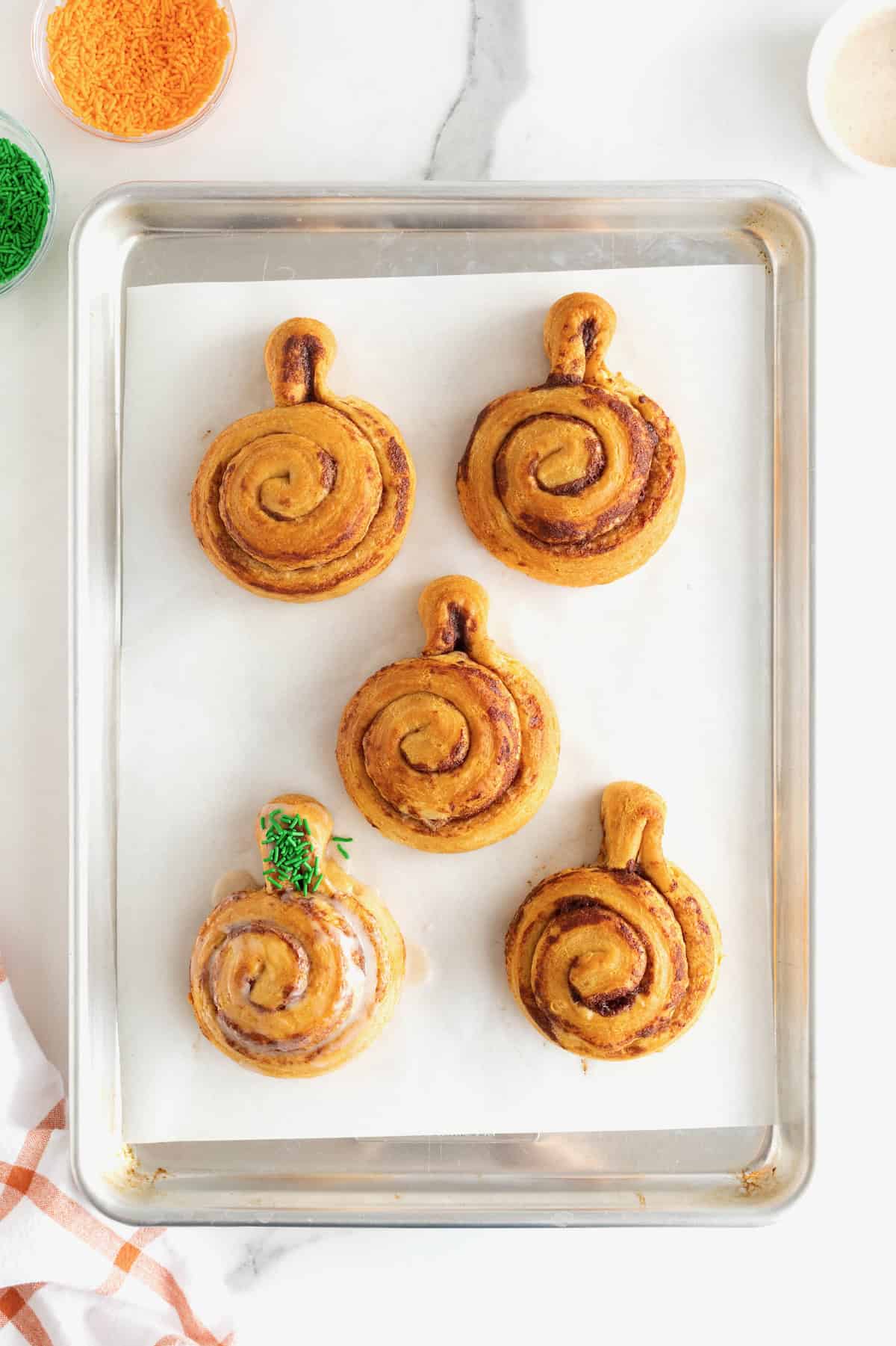 Five iced pumpkin cinnamon rolls on a parchment lined aluminum baking sheet. One of the pumpkins has  green sprinkles on the stem.