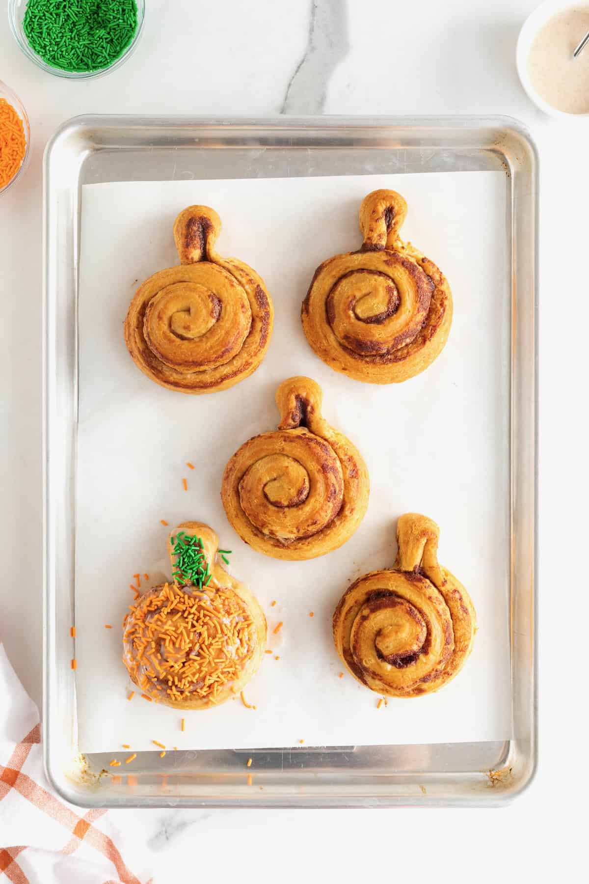 Five iced pumpkin cinnamon rolls on a parchment lined aluminum baking sheet. One of the pumpkins has orange and green sprinkles.