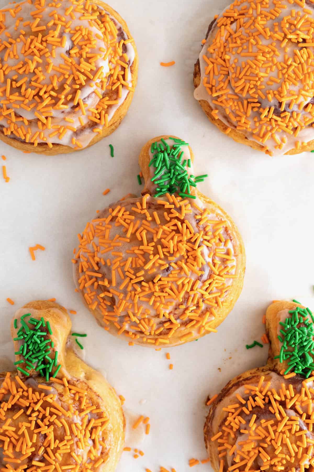 Five iced pumpkin shaped cinnamon rolls with orange and green sprinkles on a parchment lined aluminum baking sheet.