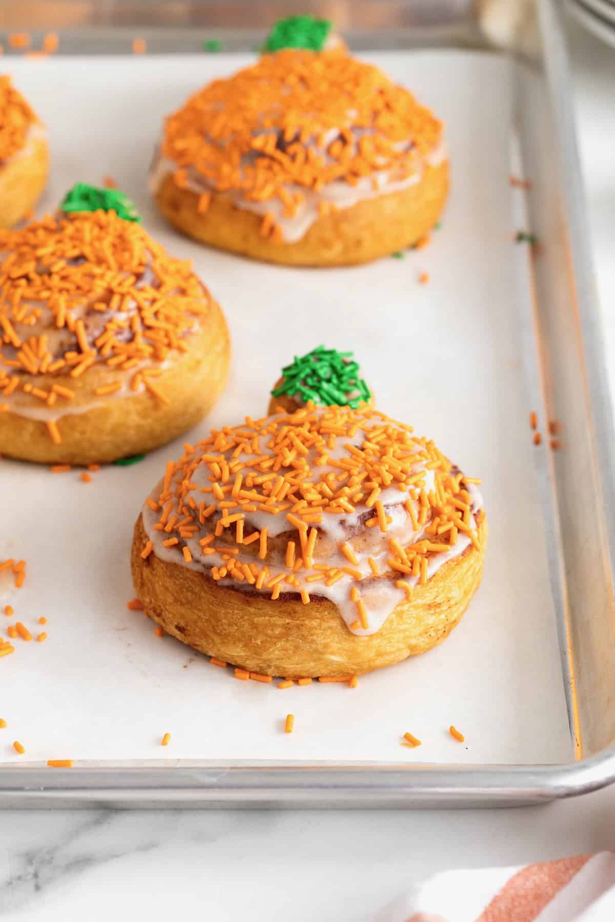 Iced pumpkin shaped cinnamon rolls with orange and green sprinkles on a parchment lined aluminum baking sheet.