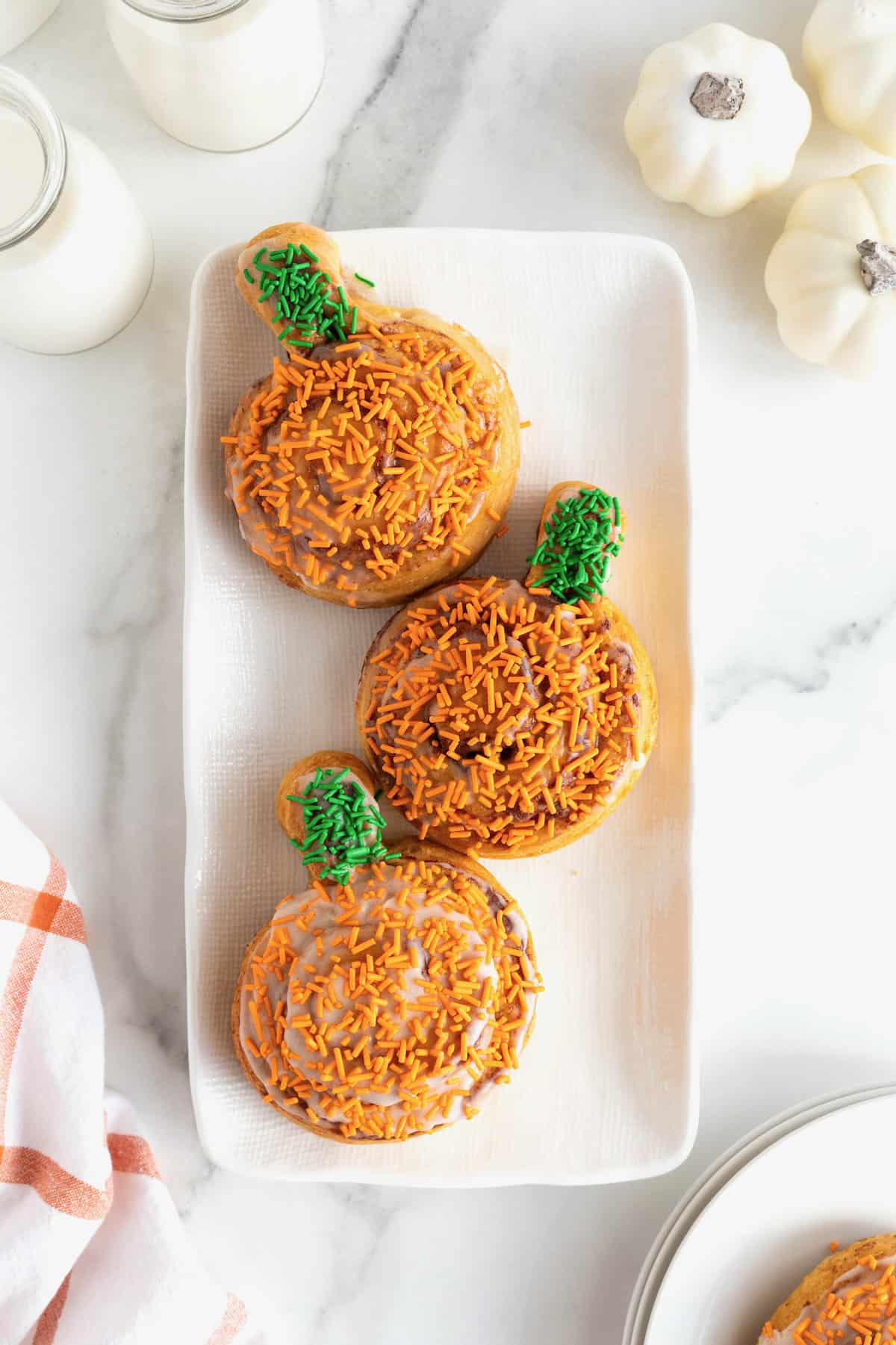 Three cinnamon rolls shaped like pumpkins with orange and green sprinkles on a rectangular white serving tray.